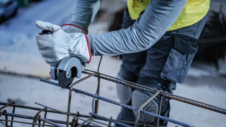 Person wearing safety equipment uses an angle grinder to cut steel rebar.