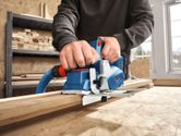 A person uses a cordless planer on a wooden board in a workshop.