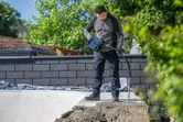 A person wearing safety equipment uses a power tool to compact concrete outdoors.