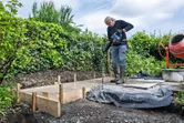 A person wearing safety equipment uses a power tool to level fresh concrete in a garden.