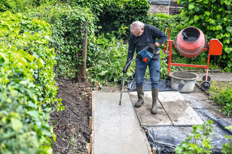 A person wearing safety equipment uses a power tool to compact concrete on a garden path.
