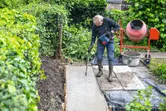 A person wearing safety equipment uses a power tool to compact concrete on a garden path.