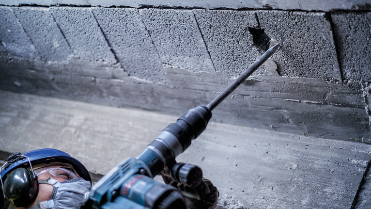 A person wearing safety equipment uses a power tool to break a hole in a concrete wall.