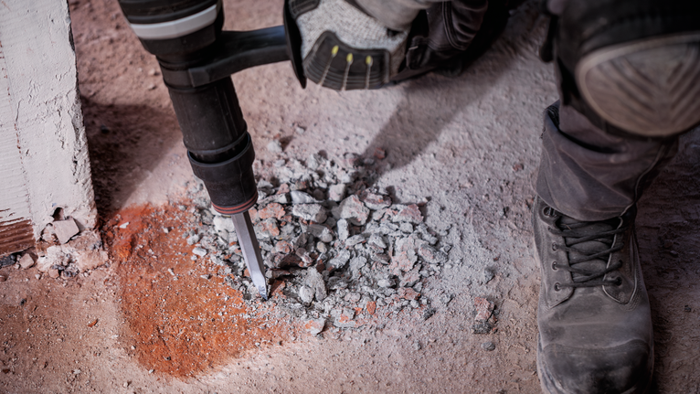 Person wearing safety equipment breaking concrete with a demolition tool.