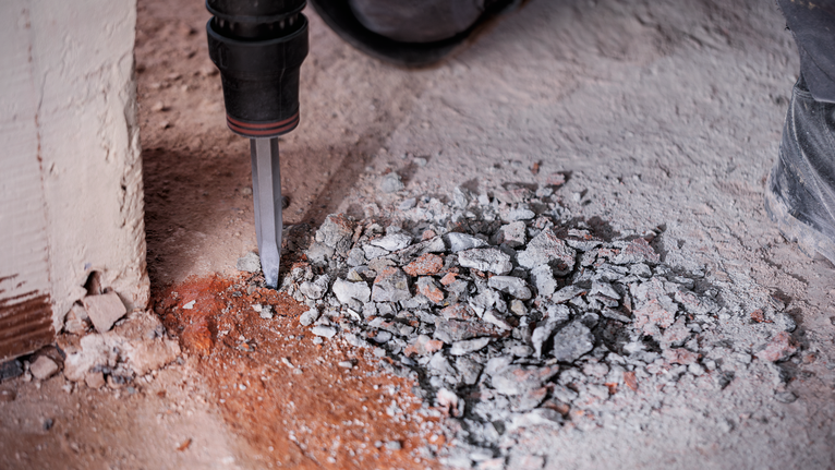 Rotary hammer breaking concrete rubble on a dusty floor.
