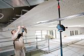 A person wearing safety equipment aligns ceiling framework using a laser leveling tool.