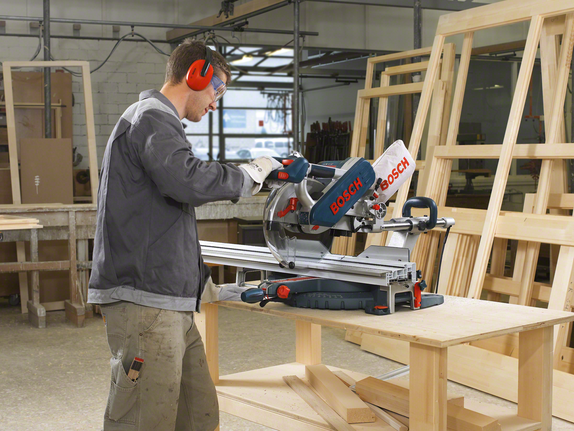 A worker wearing safety equipment cuts wood with a sliding compound miter saw.