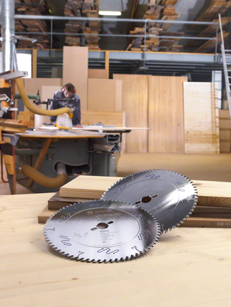Circular saw blades rest on stacked wood with a worker in the background wearing safety equipment.