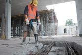 A person wearing safety equipment uses a breaker to demolish concrete at a construction site.