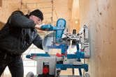 A person wearing safety equipment cuts wood with a mitre saw in a workshop.