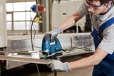 A person wearing safety equipment uses a jigsaw to cut a metal sheet.