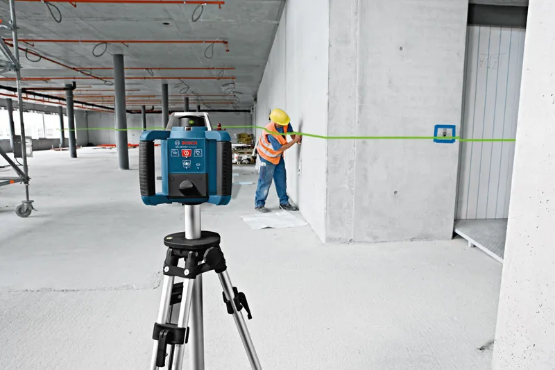 A person wearing safety equipment marks a wall using a green laser leveling tool.