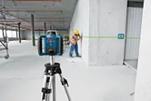 A person wearing safety equipment marks a wall using a laser leveling tool on a tripod.