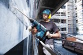 A person wearing safety equipment uses a rotary hammer to break concrete on a building site.