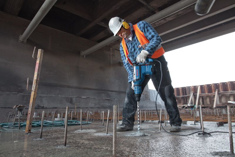 A person wearing safety equipment drills into concrete with a rotary hammer.