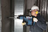 A person wearing safety equipment drills into a concrete wall using a rotary hammer.